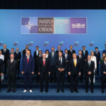 A group of world leaders in suits pose for a formal photo at the NATO Ankara Summit, standing in three rows in front of a blue backdrop with NATO and summit logos.
