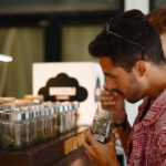 A man in a red shirt smells a jar filled with cannabis buds at a counter displaying several similar jars, while another person stands nearby.