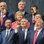 A group of world leaders from Europe in formal attire stand outdoors, looking up and pointing at something in the sky with varied expressions of curiosity and interest.