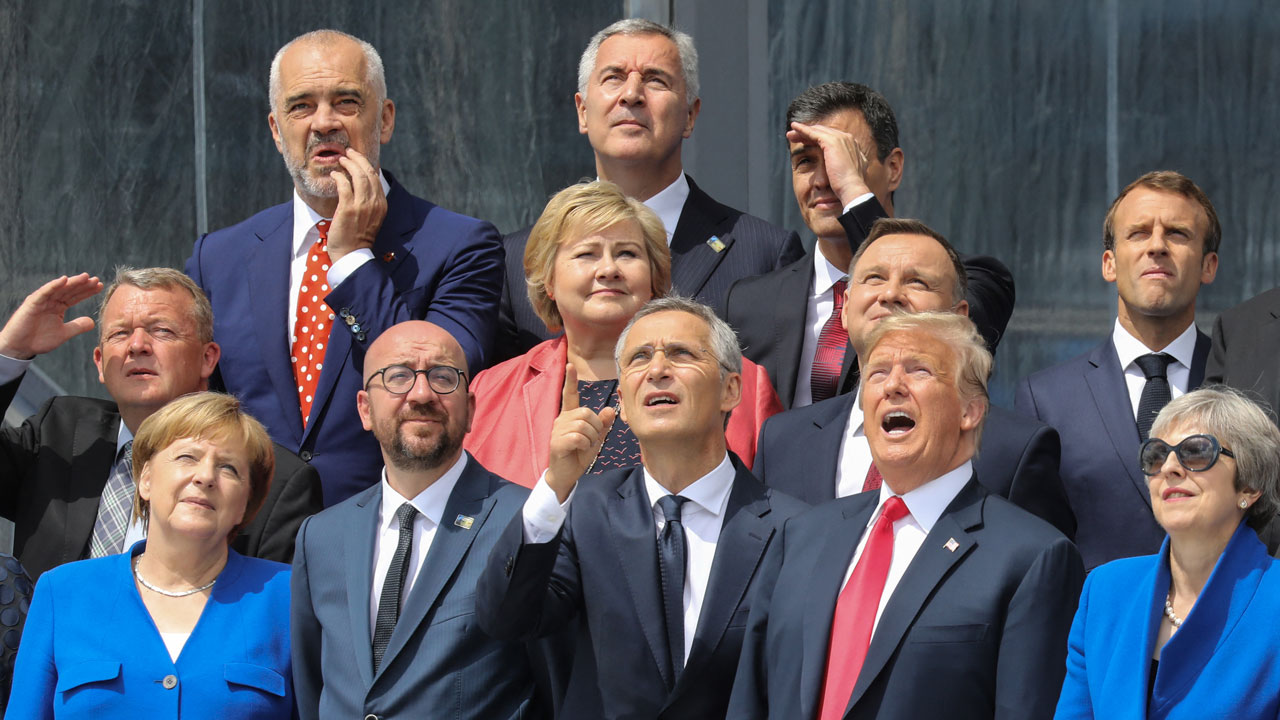 A group of world leaders from Europe in formal attire stand outdoors, looking up and pointing at something in the sky with varied expressions of curiosity and interest.