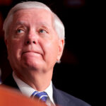 Close-up, low angle shot of Lindsey Graham looking off to his right, with a flag behind him.