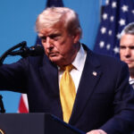 Donald Trump stands at a podium pointing, with Pete Hegseth looking on from behind and to Trump's right.