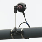 A black surveillance camera is mounted on a metal pole with brackets and a cable attached, set against a plain, light-colored background.
