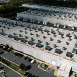 Aerial view of a large data center with rows of rooftop cooling units, surrounded by parking lots and greenery.