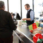 A cashier scans groceries, including bread and cheese, while a woman waits at the register in a brightly lit supermarket.