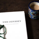 An open book titled The Odyssey lies on a table beside green glasses, inviting curiosity. A colorful mug filled with coffee or tea sits nearby, perfect for delving into the odyssey of ancient tales.