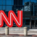 Large red CNN logo stands outside a modern glass building, reflecting nearby structures. Several concrete bollards and a sidewalk are in front of the entrance.