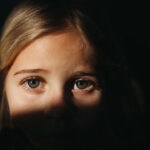 A close-up of a young child with blonde hair, partially illuminated by sunlight, with shadows obscuring the lower half of their face. The child looks directly at the camera with a serious, school-aged expression.