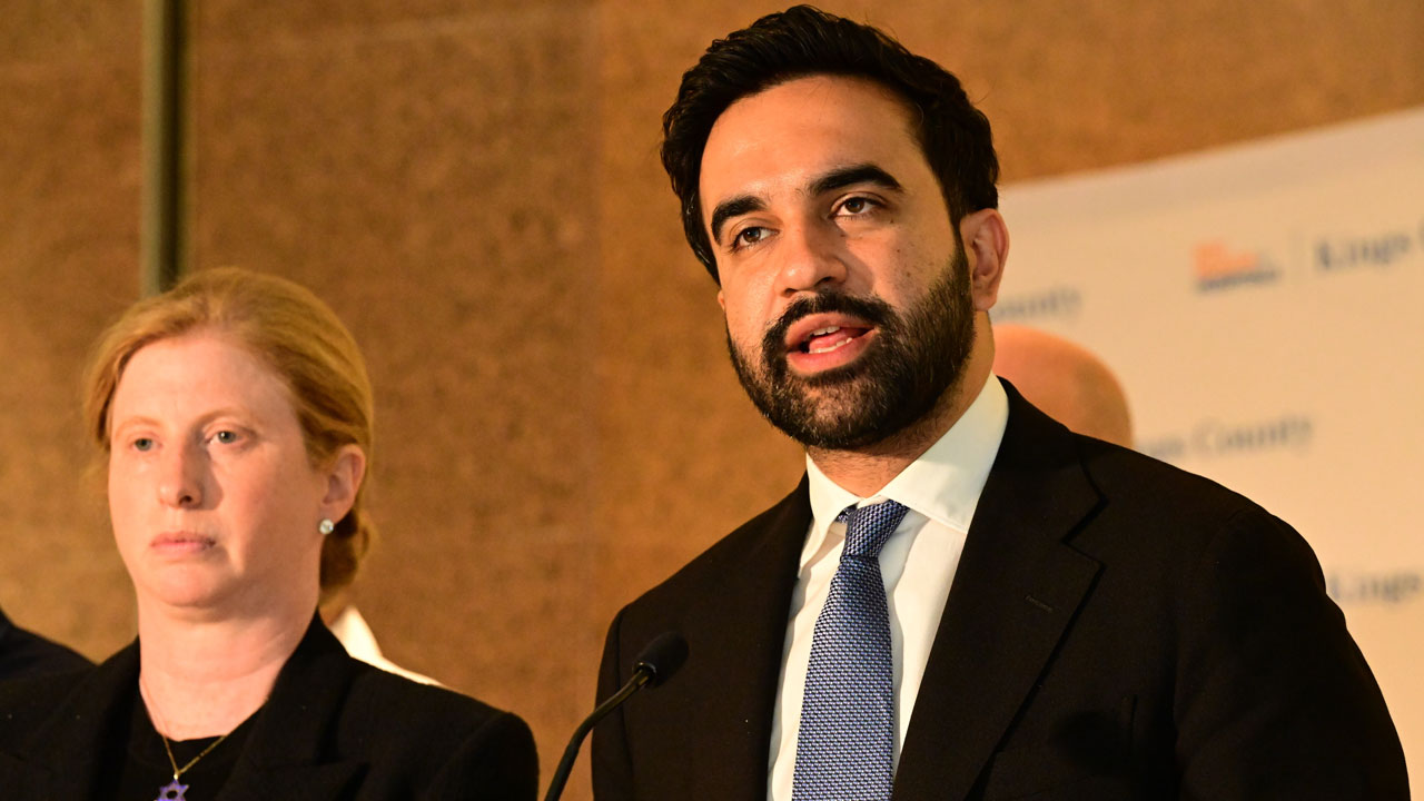 Zohran Mamdani speaks while spending next to a stone-face woman.