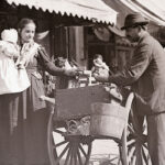 A European woman holding a baby buys something from a street vendor with a cart. Another man stands nearby. The scene takes place on a busy sidewalk in front of storefronts on a sunny day.