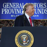 President Trump looks to his left with a smile while at a podium in front of a sign a blue and white sign reading General Motors Proving Grounds.