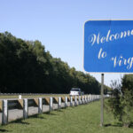 A blue Welcome to Virginia sign stands beside a highway lined with trees, greeting travelers as they drive under a clear sky—a fitting introduction to a state known for its vibrant economy.