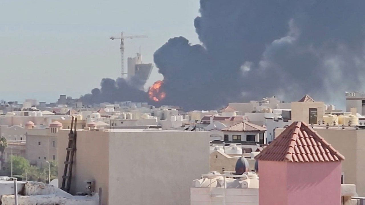 Thick black smoke and orange flames rise behind a cityscape of low, light-colored buildings; a construction crane is visible in the background.