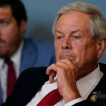Rep. Ralph Norman in a suit and red tie sits with his hand on his chin, appearing thoughtful. Another man in a blue jacket is blurred in the background.