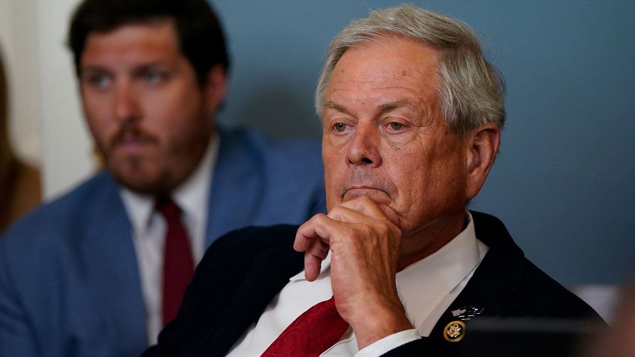 Rep. Ralph Norman in a suit and red tie sits with his hand on his chin, appearing thoughtful. Another man in a blue jacket is blurred in the background.