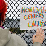A person with red and blue hair holds a colorful sign reading NO DATA CENTERS IN CALIFORNIA against a chain-link fence, protesting the expansion of data center facilities.