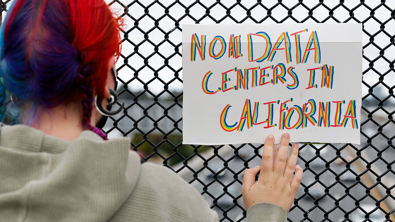 Home 3 A person with red and blue hair holds a colorful sign reading NO DATA CENTERS IN CALIFORNIA against a chain-link fence, protesting the expansion of data center facilities.
