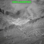 A black and white thermal or infrared image shows an explosion in a mountainous area, with the word UNCLASSIFIED in green at the top. Dust and smoke rise from the explosion among steep cliffs.
