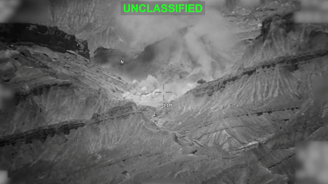 A black and white thermal or infrared image shows an explosion in a mountainous area, with the word UNCLASSIFIED in green at the top. Dust and smoke rise from the explosion among steep cliffs.