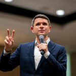 James Talarico in a dark suit holds a microphone with left hand and holds up two fingers with his right.