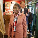 Winsome Earle-Sears in a pink suit with red, white, and blue scarf enters an event trailed by Glenn Youngkin.