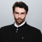 Hasan Piker in a black button-up jacket against a grey background.