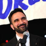 Zohran Mamdani smiling while at a microphone.