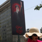 A large mural of the late Iranian Ayatollah Khamenei raising his fist is displayed on the side of a building. A woman in a wide-brimmed hat and sunglasses walks in the foreground. Trees and other buildings are visible nearby.