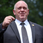 Tom Homan in a suit and patterned tie gestures with his right hand while speaking outdoors, with green trees blurred in the background.