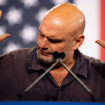 John Fetterman raises both hands while speaking at a podium, with a blurred U.S. flag in the background. Tattoos are visible on his left forearm, and he is wearing a dark shirt.