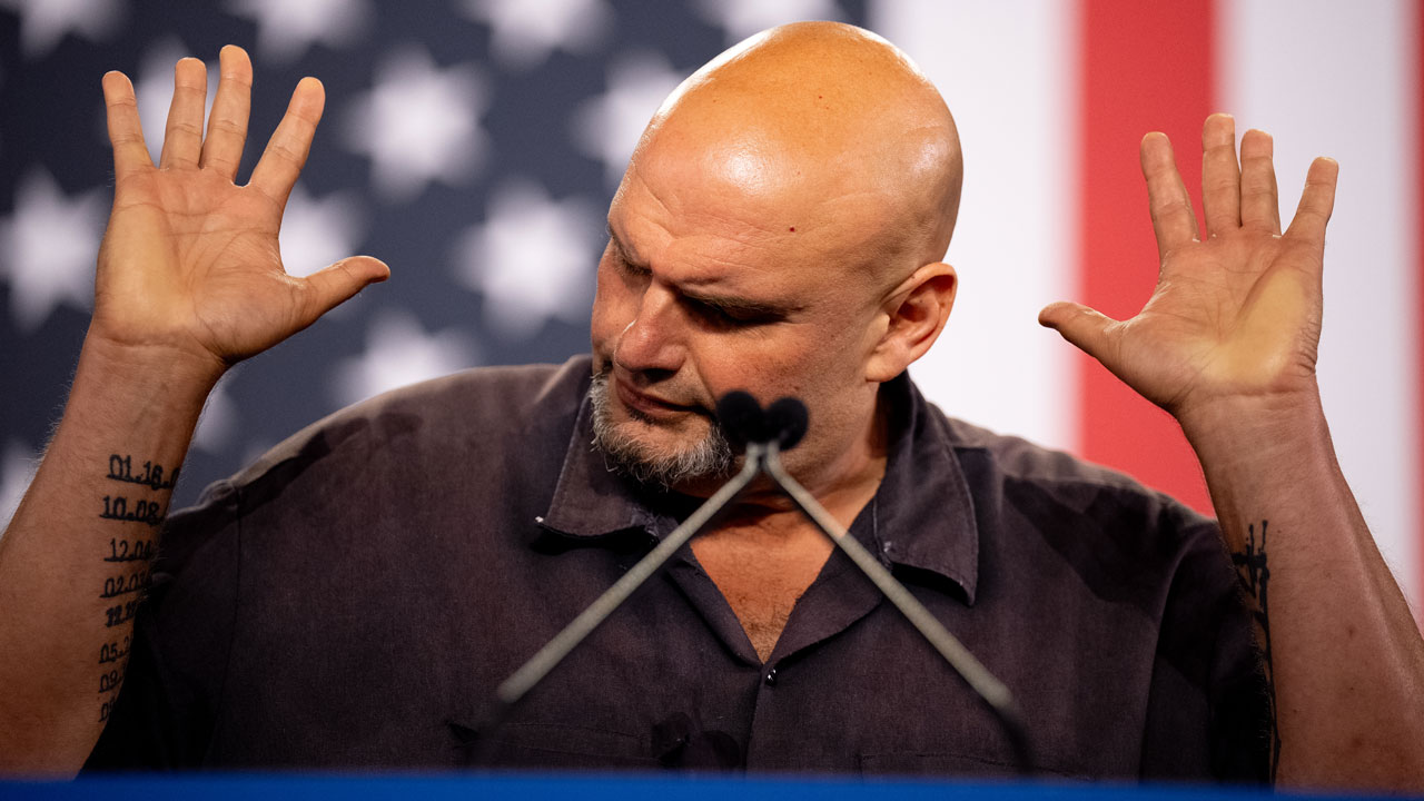 John Fetterman raises both hands while speaking at a podium, with a blurred U.S. flag in the background. Tattoos are visible on his left forearm, and he is wearing a dark shirt.