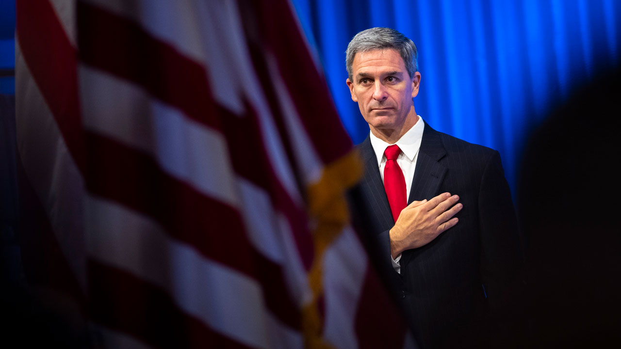 Ken Cuccinellil in a suit with a red tie stands with his hand over his heart, looking forward. An out-of-focus American flag is in the foreground, and blue curtains are in the background.