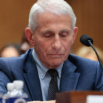 Dr. Anthony Fauci in a blue suit and tie, sits at a desk with a microphone, looking down. There are water bottles in the foreground and blurred people in the background.