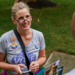 Judi Briskman, with glasses and campaign buttons stands outside on grass, talking to another person. She holds pamphlets, wears a gray When We Vote shirt, and smiles while listening.