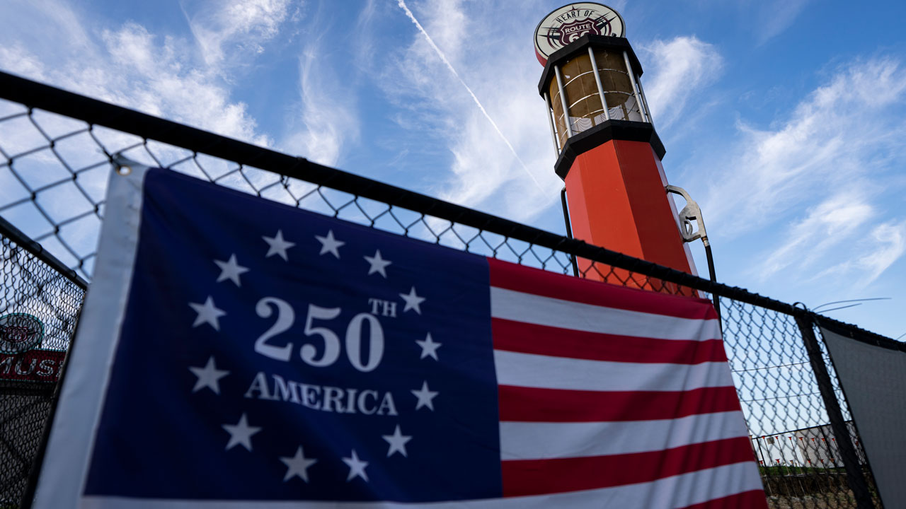 A flag reading America 250 hangs on a chain-link fence, with a red and white tower in the background under a blue sky with scattered clouds and a contrail.