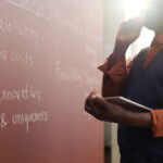 Person in a vest holds a tablet while thinking and looking at notes written in chalk on a red wall, with sunlight shining in from the side—a moment that reflects the thoughtful analysis often found in higher education settings.