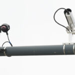 Two surveillance cameras, resembling a watchful flock, are mounted on a cylindrical metal pole, with cables connected to each camera, against a plain white background.
