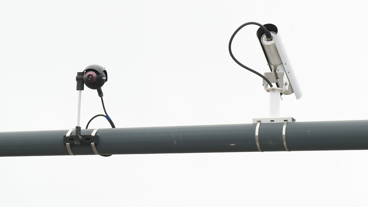 Two surveillance cameras, resembling a watchful flock, are mounted on a cylindrical metal pole, with cables connected to each camera, against a plain white background.
