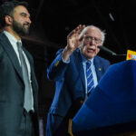 Sen. Bernie Sanders speaks at a podium labeled 100 Day Address beside Zohran Mamdani in a gray suit, under indoor lighting.