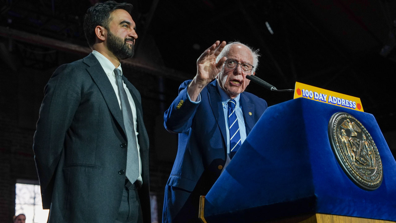 Sen. Bernie Sanders speaks at a podium labeled 100 Day Address beside Zohran Mamdani in a gray suit, under indoor lighting.