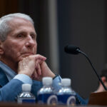 Dr. Anthony Fauci sits at a desk with his hands clasped, listening thoughtfully. A microphone and water bottles are in front of him, and several people are blurred in the background.