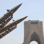 Three military missiles are displayed in front of the Azadi Tower in Tehran, Iran, under a clear blue sky. The image captures both the missiles and the iconic Iranian landmark from a low angle, highlighting Iran’s blend of modern military presence and historic architecture.