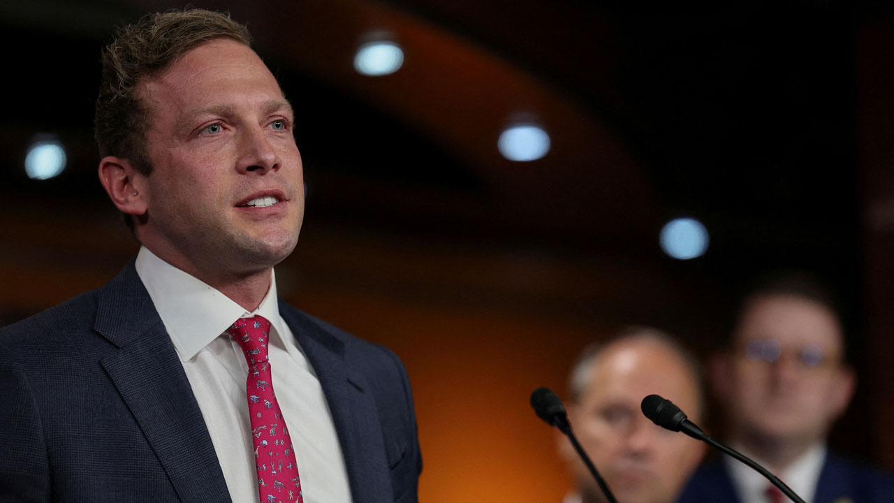Home 1 Rep. Max Miller in a suit and red tie speaks at a podium, with two blurred people in the background under warm lights.
