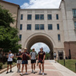 A group of students walk on a pathway toward a large Texas campus building with an archway, surrounded by trees and a partly cloudy sky.