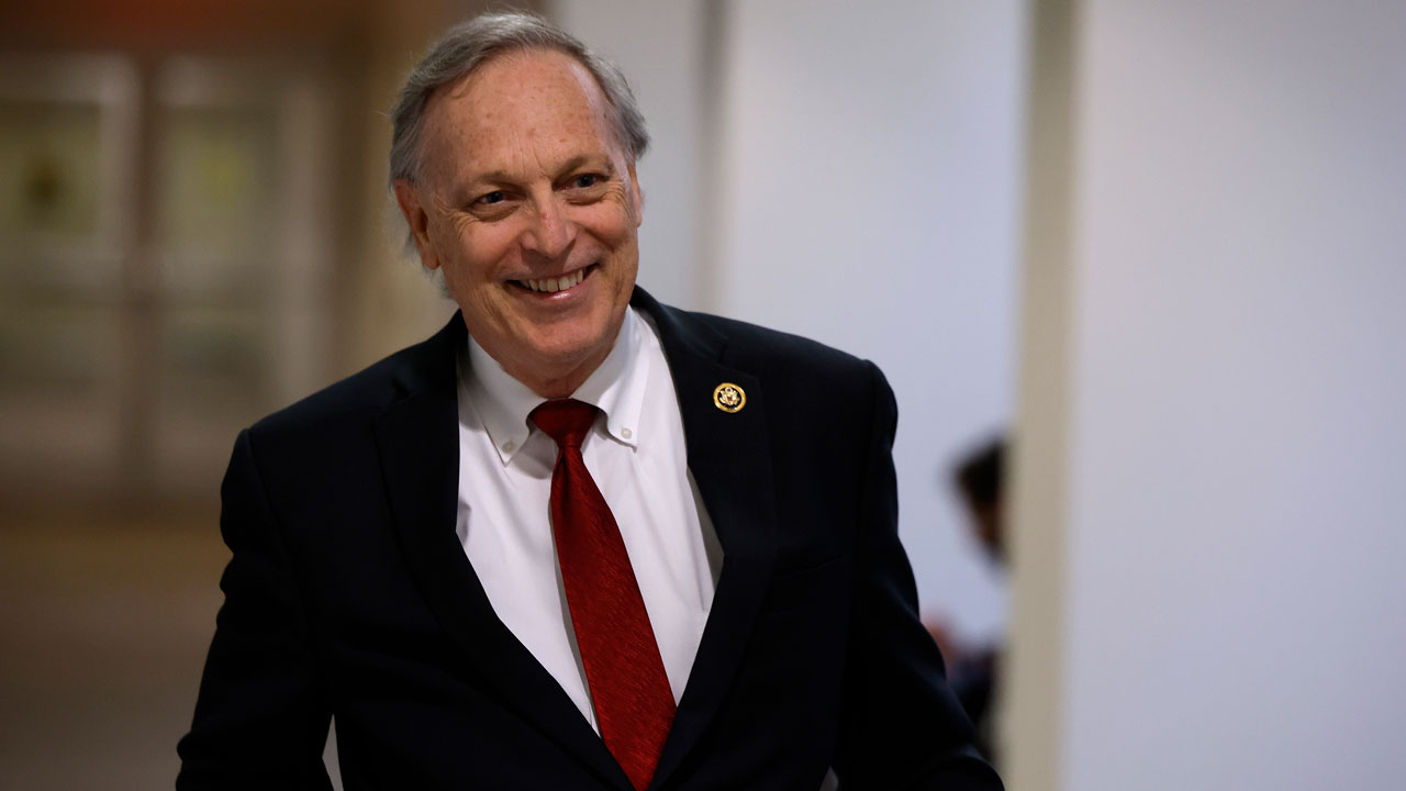 Rep. Andy Biggs in a dark suit, white shirt, and red tie walks down a hallway with a blurred background.