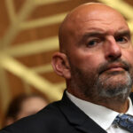 John Fetterman, wearing a suit and patterned tie, sits in front of a geometric gold background, looking slightly to the side with a serious expression.
