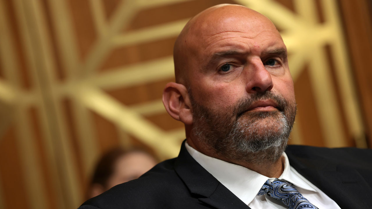 John Fetterman, wearing a suit and patterned tie, sits in front of a geometric gold background, looking slightly to the side with a serious expression.
