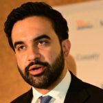 Zohran Mamdani, wearing a suit and tie, speaks in front of a blurred background with indistinct text and logos.