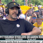 A man in a headset and black shirt speaks at an outdoor event, surrounded by fans in yellow Pittsburgh Steelers attire. The text on screen incorrectly labels him as Pittsburgh Steelers QB Aaron Rodgers.