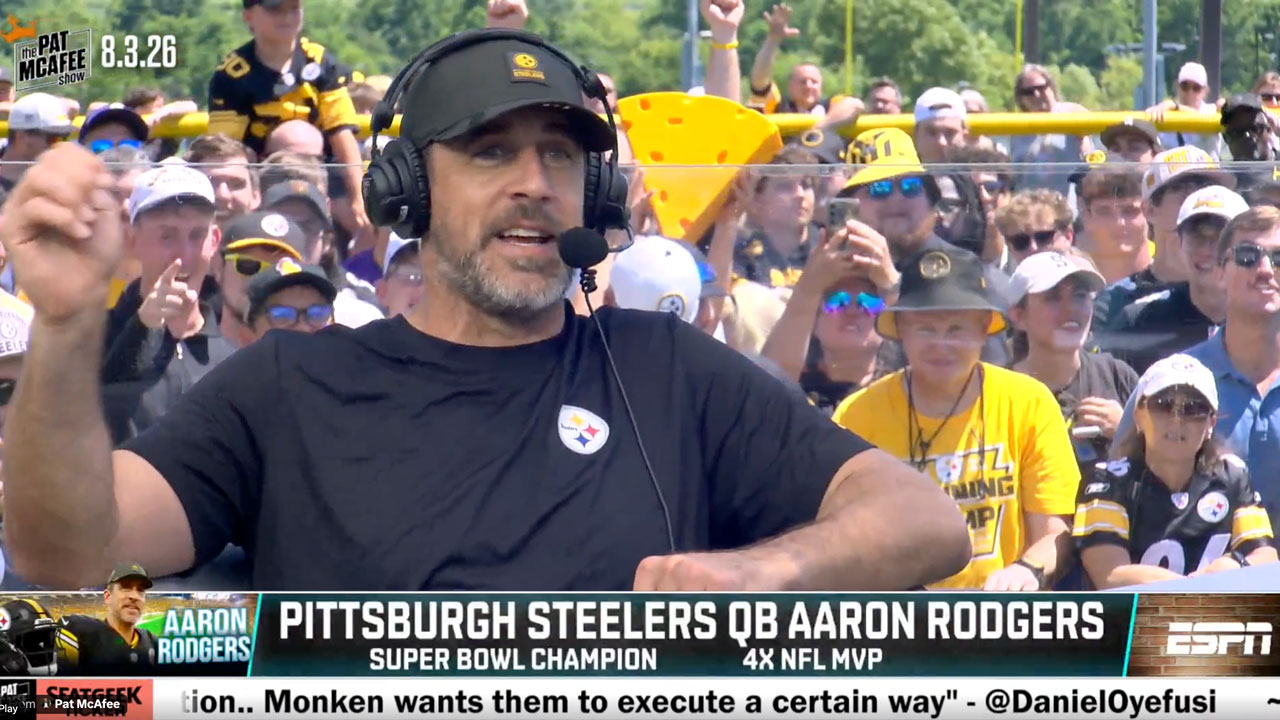 A man in a headset and black shirt speaks at an outdoor event, surrounded by fans in yellow Pittsburgh Steelers attire. The text on screen incorrectly labels him as Pittsburgh Steelers QB Aaron Rodgers.